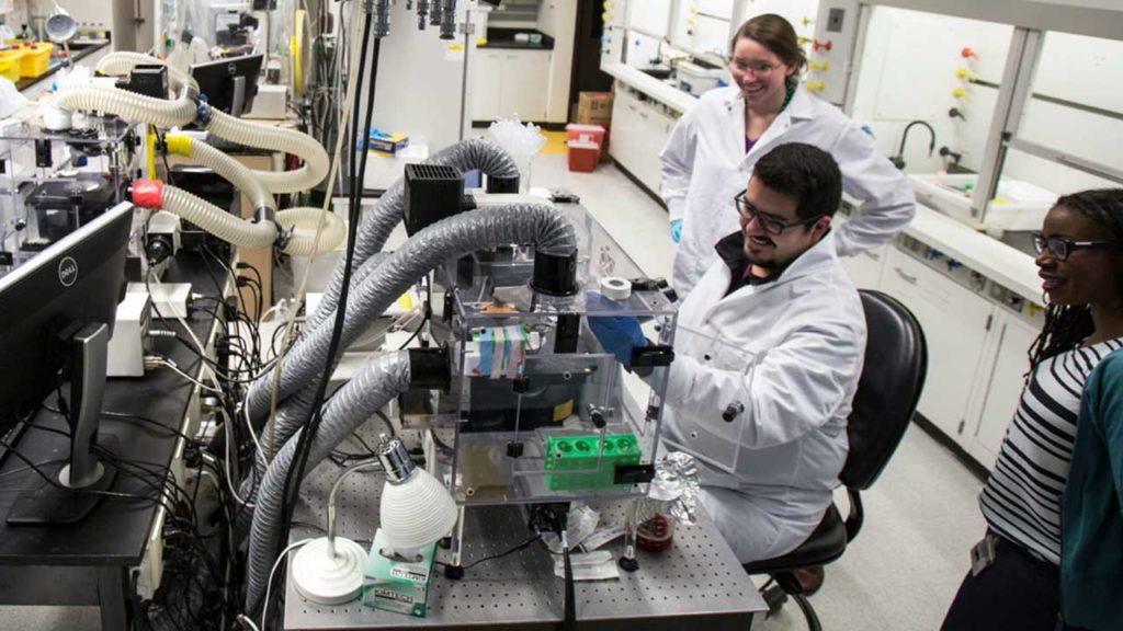 Three people in lab coats working with laboratory equipment at a workstation in a lab.