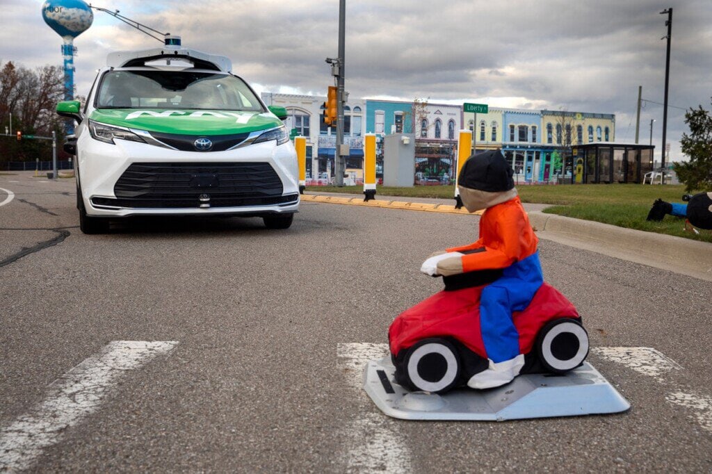 During an autonomous vehicle test at M city, a mannequin that represents a child on a riding toy is situated in the street in a crosswalk as a May Mobility AV approaches. In the background are the mock building facades of M city's downtown.