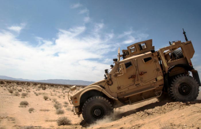 Side view of a khaki-colored military ATV in the desert on a partially cloudy day. Its front tire kicks up a puff of dust as it offroads down a rocky hill.
