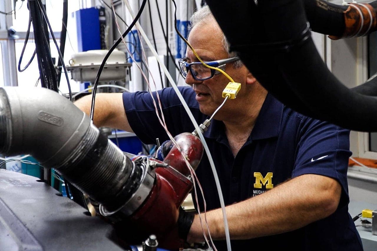 Wires hang from above and an industrial metal tube just bigger than an adult arm juts out from the left in this image of mechanical engineering professor Andre Boehman working on a heavy duty single cylinder engine in the University of Michigan's Auto Lab. Boehman wears a blue U-M polo shirt and protective goggles. 