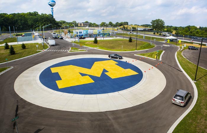 An aerial view of the Mcity test facility's roundabout that features a giant University of Michigan maize-colored block M on a blue background in the center. A vehicle navigates the roundabout. In the background, patches of green grass represent medians and a city square outside Mcity's mock downtown. 