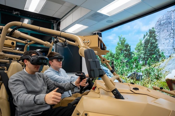 Two male students sit in a military utility vehicle simulator wearing virtual reality goggles and holding control sticks. 
