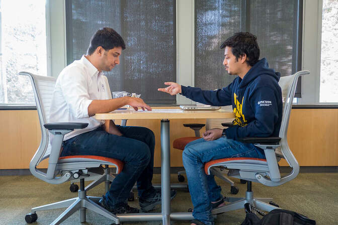 Two men sit facing each other at a table discussing. 